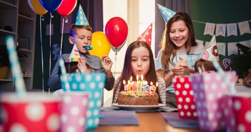 Une petite fille heureuse fête son anniversaire avec ses amis à la maison, souffle les bougies sur le gâteau, fait un vœu alors qu'elle est assise à la table du salon et que les enfants applaudissent et rient.