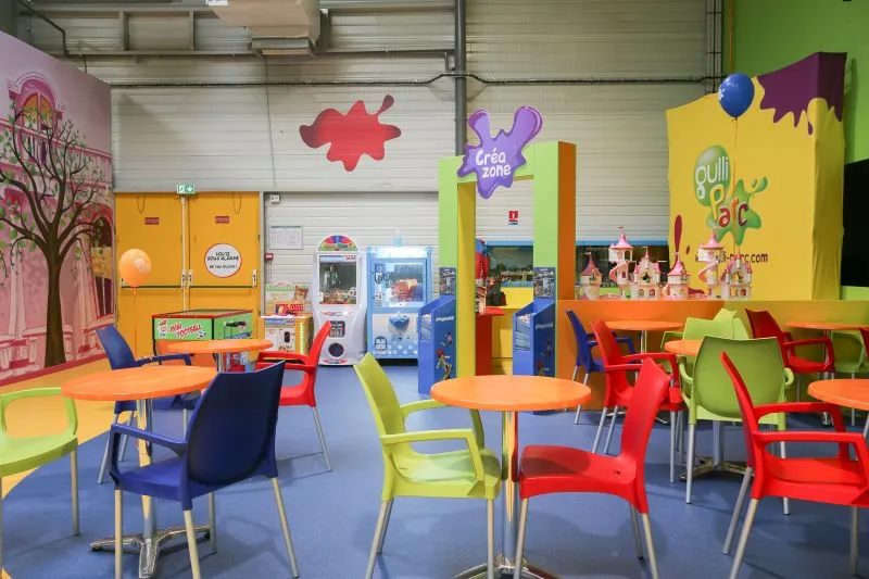 L'image montre une salle de Gulli Parc avec des tables et des chaises colorées, ainsi que des jeux au fond de la salle.