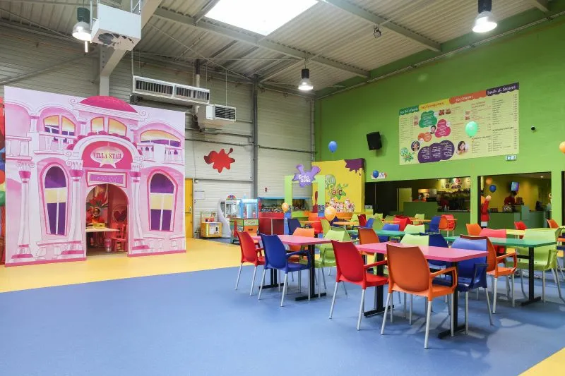 L'image montre une salle de Gulli Parc avec des tables et des chaises colorées, ainsi que des jeux au fond de la salle.